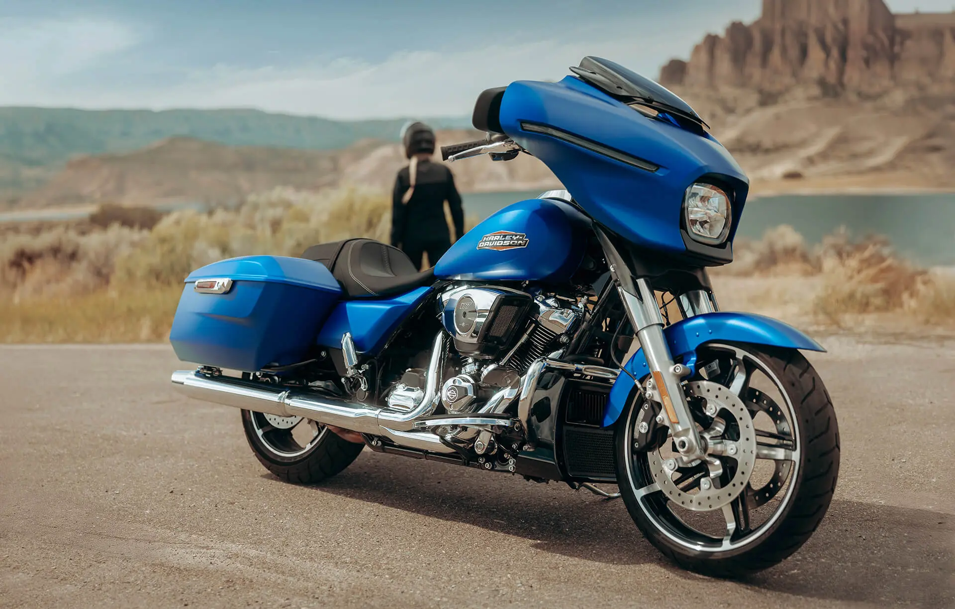 A bright blue Harley-Davidson Street Glide motorcycle with a Batwing fairing and hard saddlebags, parked on a roadside with a person and the Grand Canyon in the background